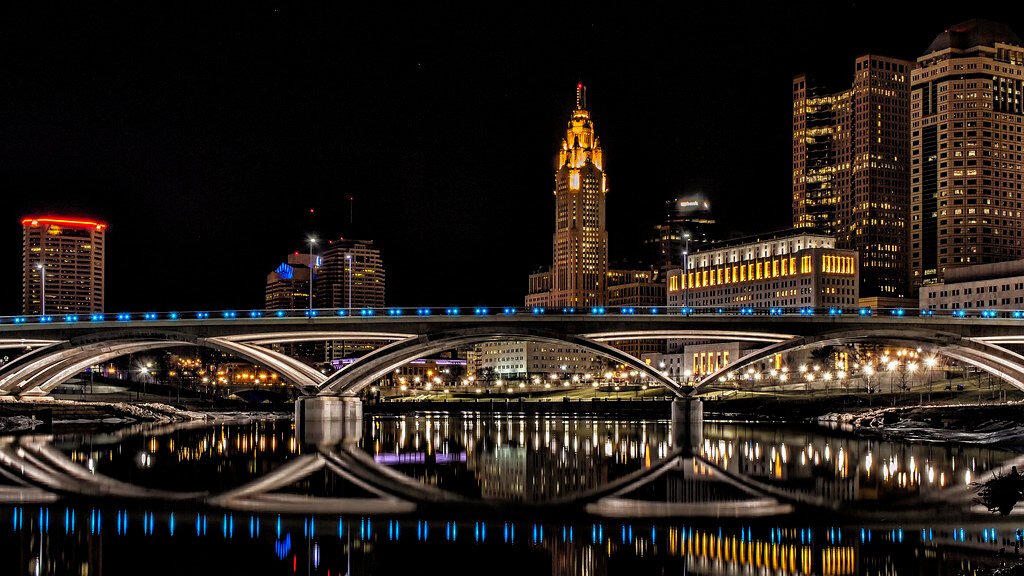 A stunning night view of a bridge over a river, with the city skyline beautifully mirrored in the calm water. Pest control in Columbus keeps the city clean.